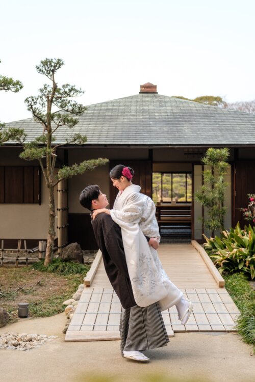 Groom in montsuki hakama giving the bride in shiromuku a princess carry on a stone path in a Japanese garden, kimono wedding photos in Japan