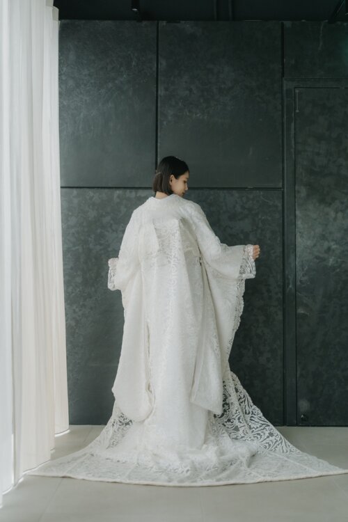 Back view of a bride in a shiromuku trailing its long lace-like hem between white curtains and a dark wall, kimono wedding photos in Japan (DE101).