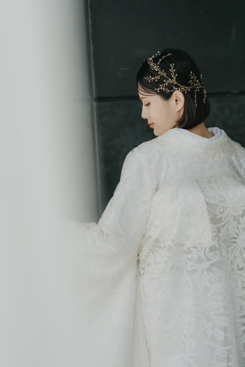 Bride in a shiromuku looking down over her shoulder, a gold branch headpiece and embroidered floral pattern visible in this back view, kimono wedding photos in Japan (DE101).