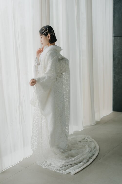 Side view of a bride in a shiromuku with a long lace-like hem by a window draped in white curtains, natural-light studio wedding photography in Japan (Shiromuku DE101).