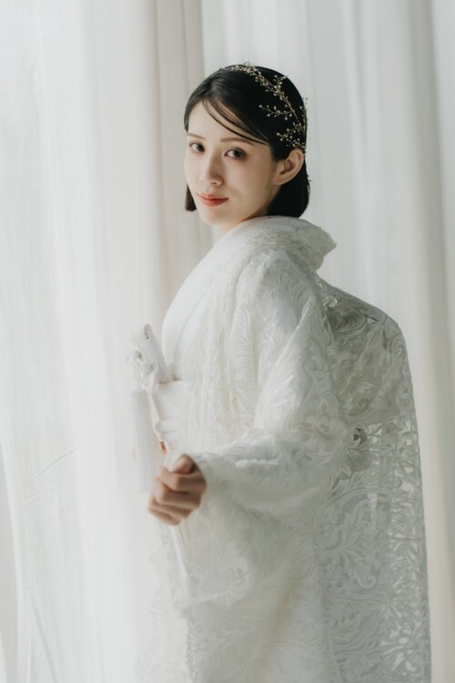 Bride in a shiromuku holding a fan to her chest and gazing at the camera in soft curtain-filtered light, a pre-wedding photoshoot in Japan (Shiromuku DE101).