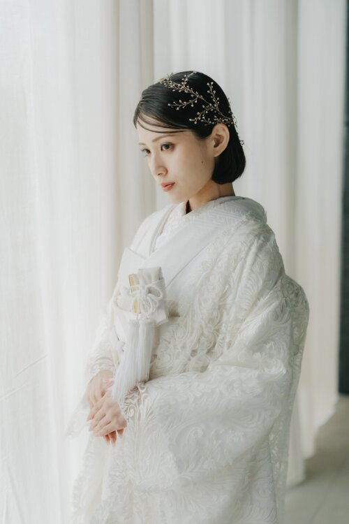 Bride in a shiromuku with hands folded and eyes lowered before white curtains, her gold headpiece pairing with the lace-like pattern in this Japan wedding photography portrait (DE101).