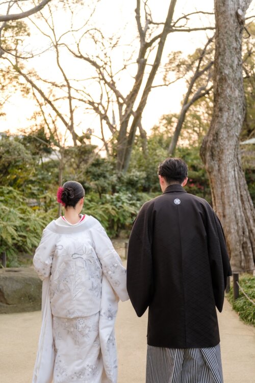 Back view of a bride in shiromuku and a groom in black montsuki hakama standing side by side in a Japanese garden at dusk, a pre-wedding photoshoot in Japan