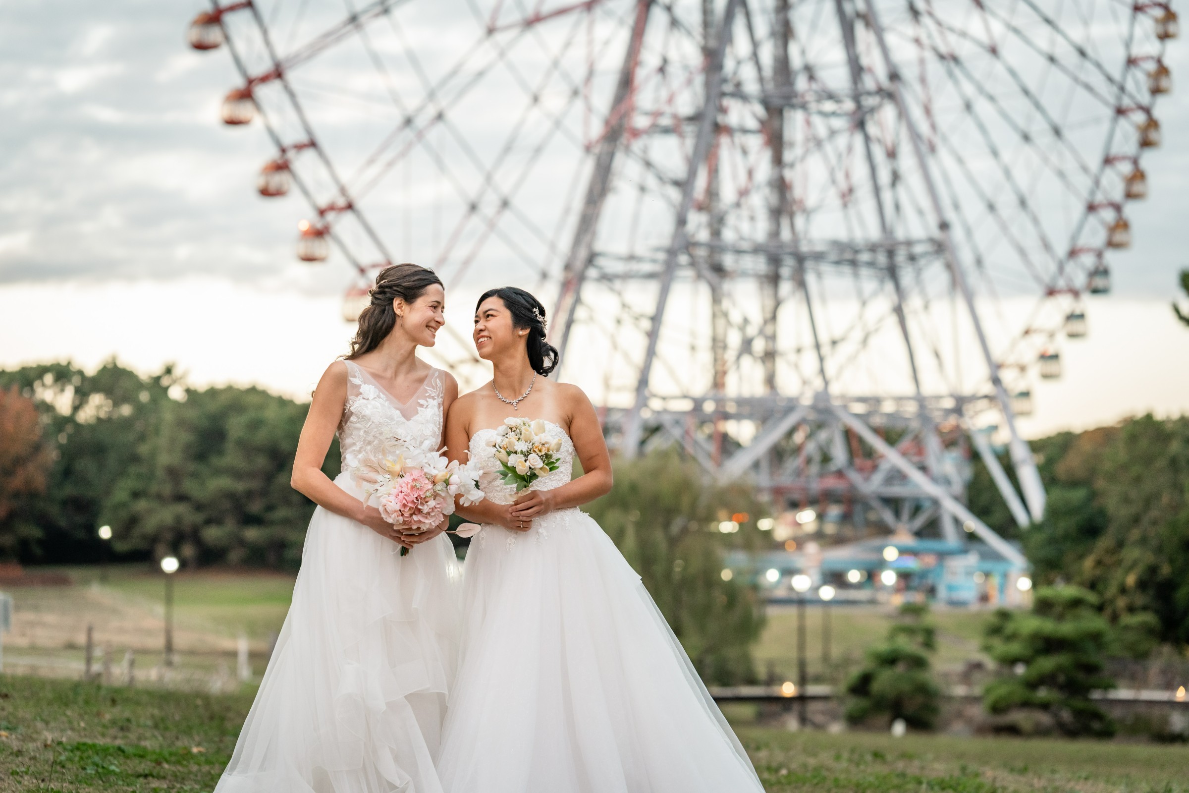 Two brides in white gowns smiling at each other, holding bouquets, with a large Ferris wheel rising behind them at dusk.