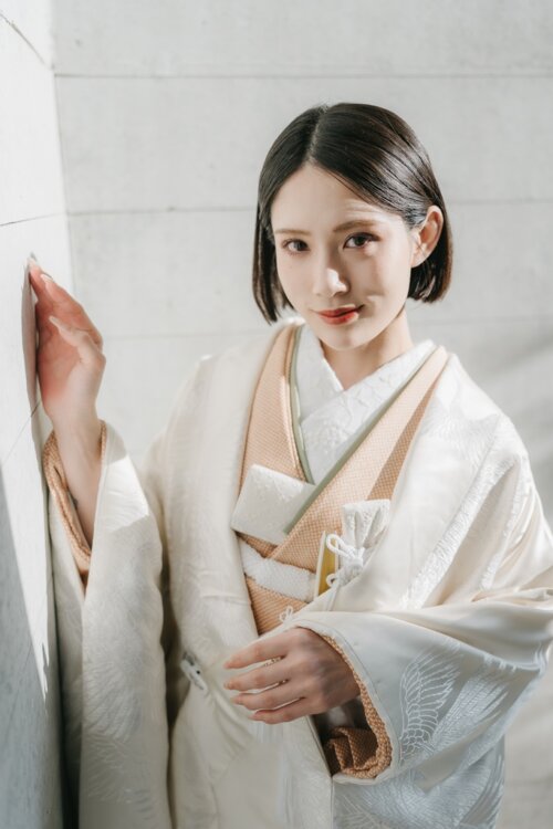 Bride in a shiromuku with a beige underkimono resting a hand on a white wall and gazing at the camera, the crane pattern shining in this studio wedding photo in Japan