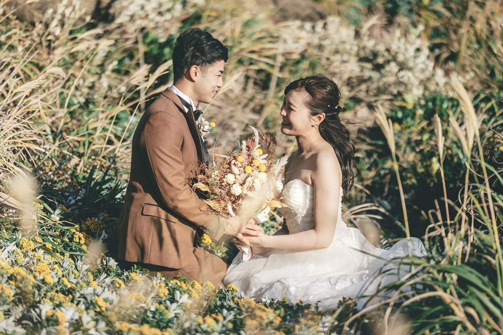 Bride in a wedding dress and groom in a brown suit sitting together among pampas grass on Jogashima, autumn wedding photos near Tokyo