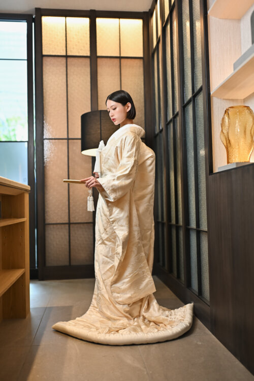 Bride in a shiromuku turning sideways with a fan and trailing hem in a modern Japanese room lit by latticed windows, the pattern on the back visible in this pre-wedding photoshoot in Japan (Shiromuku AB102).