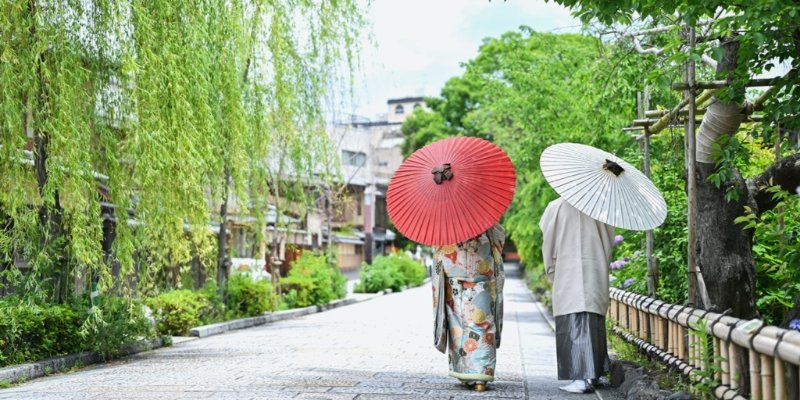A couple walks a willow-lined street holding red and white parasols, representing the Wedding Avenue official Japanese site.