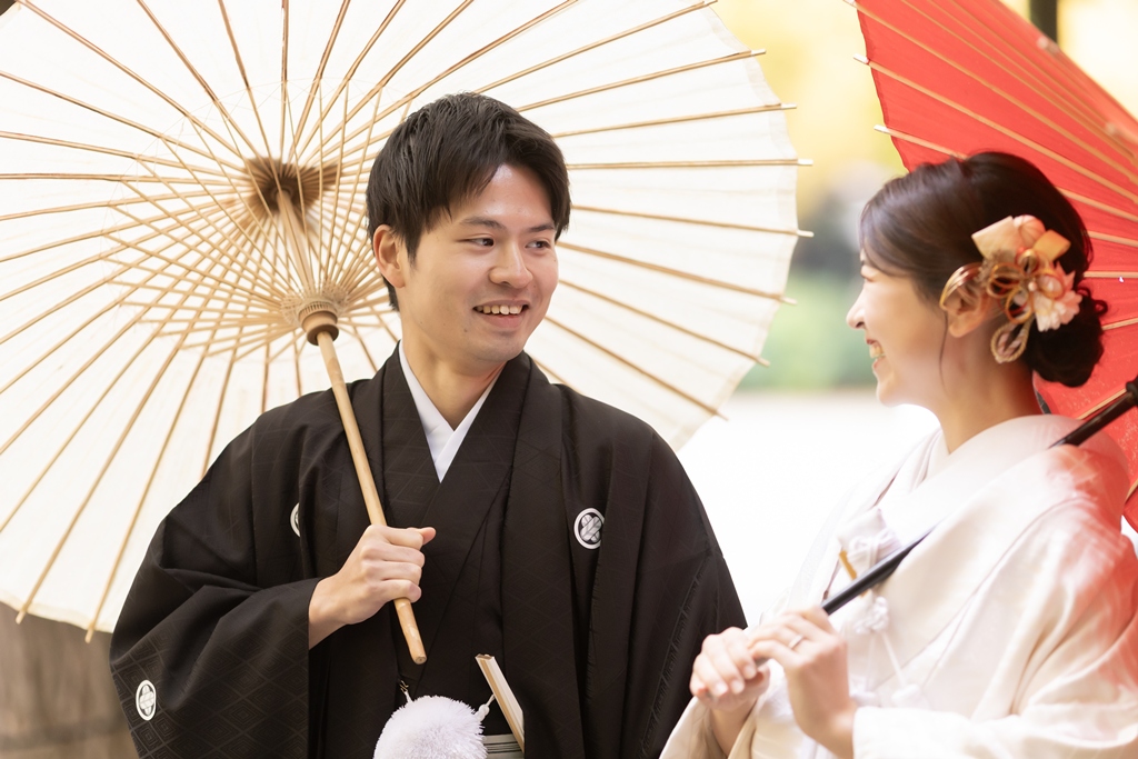 Bride in shiromuku and groom in montsuki laughing together under red and white Japanese umbrellas at the Jingu Gaien Ginkgo Avenue