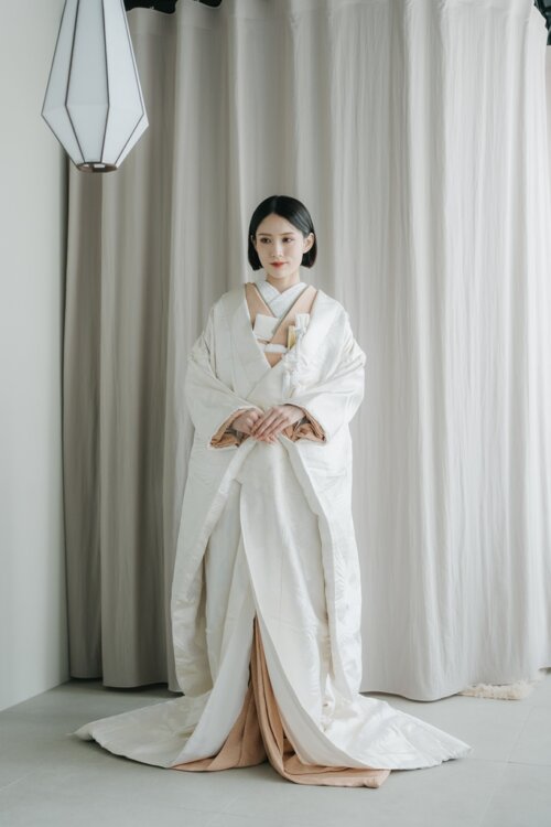 Bride in a shiromuku white kimono standing with hands folded and the hem spread out under pendant lights, a front view from a studio kimono wedding photoshoot in Japan