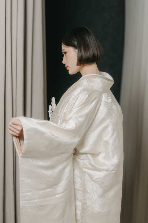 Bride in a shiromuku spreading her sleeve and showing her profile before a curtain, the woven crane pattern visible in this studio Japan wedding photography shot (DR101).