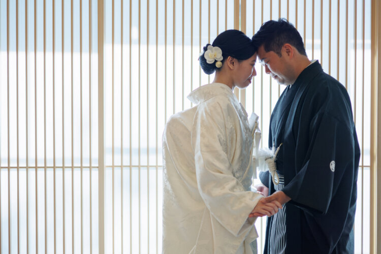 Bride in shiromuku with a white orchid in her hair and groom in montsuki hakama hold hands with foreheads touching by the lattice screens, kimono wedding photos