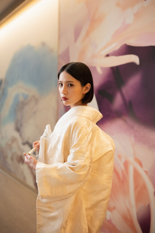 Bride in a shiromuku glancing back with a fan against a pink and purple floral art wall, a vivid pose for kimono wedding photos in Japan (AB102).