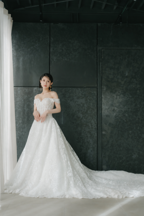 Bride in an off-shoulder all-lace wedding dress standing between a dark stone-textured wall and white curtains during a studio wedding photoshoot in Japan