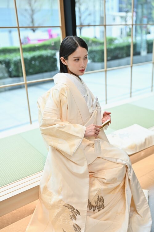 Bride in a shiromuku seated by a window on a raised tatami platform, fan in hand and gazing outside, the gold feather hem shining in this kimono wedding photo in Japan (Shiromuku AB101).