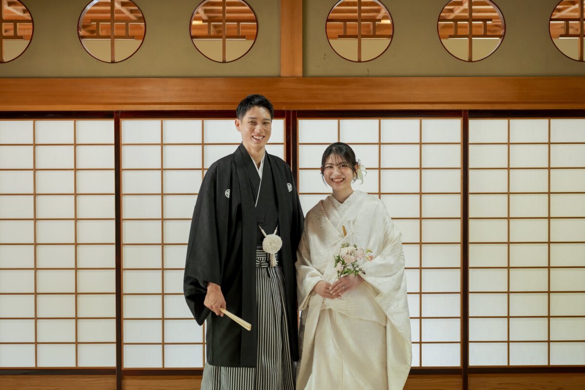 Groom in montsuki holding a folding fan and bride in shiromuku holding a pale pink bouquet in front of shoji screens and round lattice windows, a classic kimono wedding photo pose