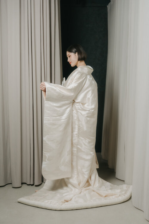 Bride in a shiromuku looking down and spreading her sleeves and long hem before a gray curtain, a full-length kimono wedding photo in Japan (Shiromuku DR101).