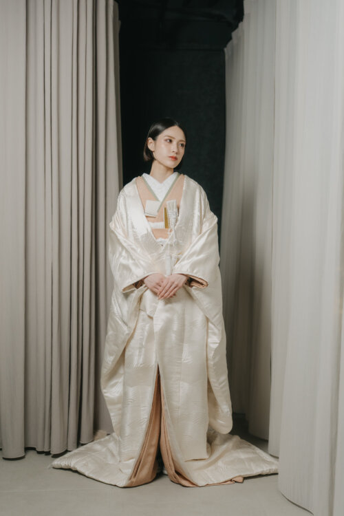 Bride in a shiromuku standing front-facing with hands folded before a curtain, the spread hem giving an elegant full-length look in this Japan wedding photography shot (Shiromuku DR101).