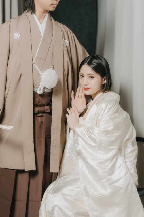 Bride in a shiromuku seated at the feet of the groom in a beige montsuki and hakama, hand to her cheek and gazing at the camera, a pre-wedding photoshoot in Japan (DR101).