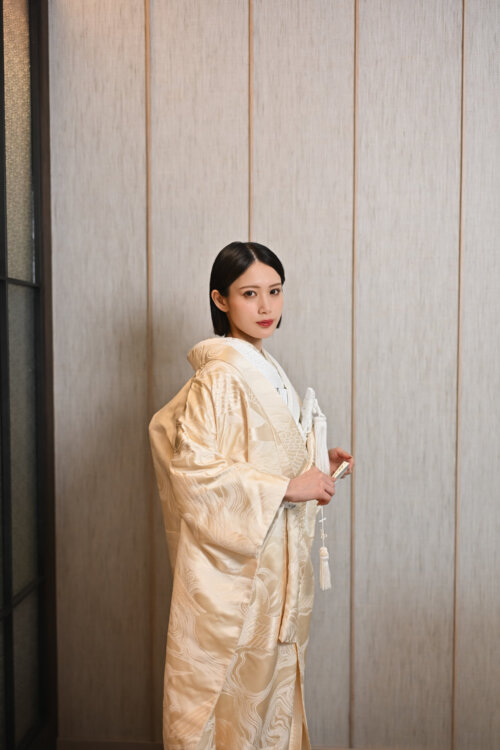 Bride in a shiromuku with gold embroidery standing front-facing with a fan and tassels before a wooden wall, Japan wedding photography from our studio (AB102).