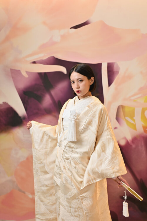 Bride in a gold crane shiromuku spreading her sleeves with a fan before a large floral art wall, a colorful studio backdrop for a pre-wedding photoshoot in Japan (Shiromuku AB102).