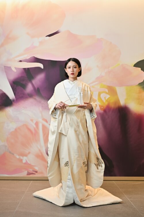 Bride in a shiromuku standing front-facing with a fan and spread hem before a floral art wall, the feather pattern on the hem visible in this Japan wedding photography shot (AB101).