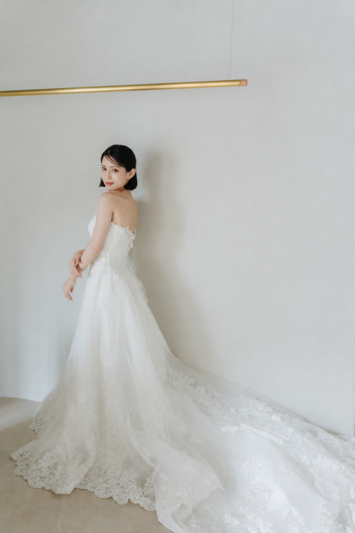 Bride looking back in front of a white wall as the long lace train of her wedding dress spreads out, a back view from a pre-wedding photoshoot in Japan