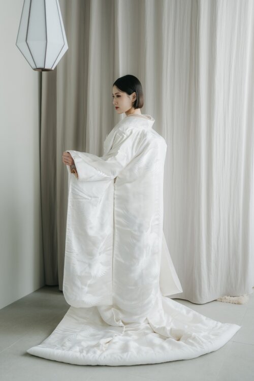Bride holding her sleeve and trailing the long hem of her shiromuku in profile before a curtain, a full-length silhouette from a pre-wedding photoshoot in Japan