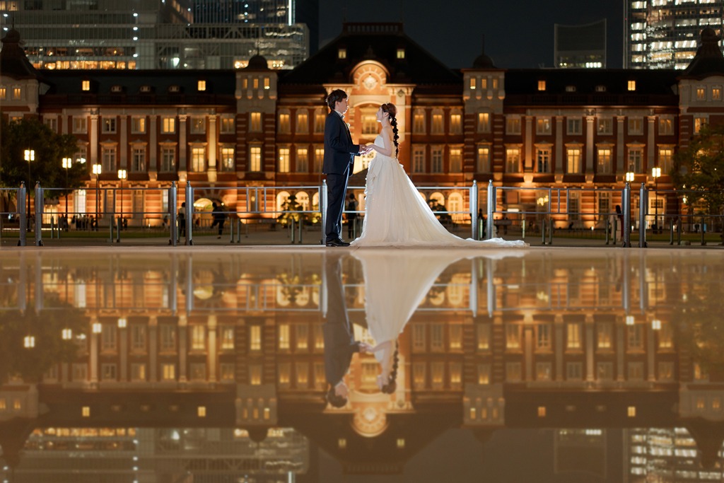 A couple facing each other as Tokyo Station at night is mirrored in the water, a reflection shot from a Japan wedding photography and movie plan