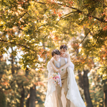 Groom giving the bride in a wedding dress a piggyback ride under trees in autumn colors, both laughing during a fall foliage pre-wedding photoshoot in Japan