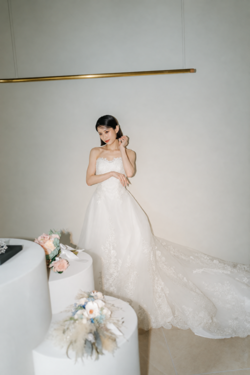 Bride in the A-line DC08 wedding dress posing with a hand by her ear beside a flower-topped column, a studio shot for Japan wedding photography