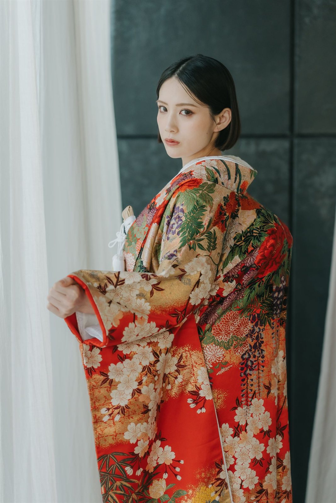 Bride glancing back in a red iro-uchikake kimono woven with cherry blossoms and peonies, soft window light, a modern look for kimono wedding photos in Japan.