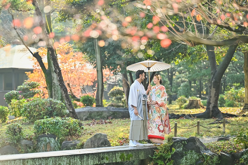 Bride in iro-uchikake and groom in hakama sharing a white umbrella on a stone bridge under autumn leaves, a kimono pre-wedding photoshoot in Osaka