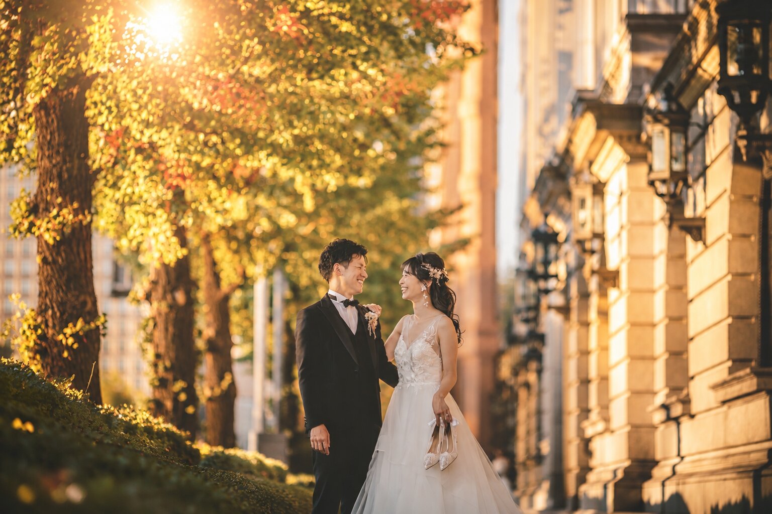 Bride in a wedding dress and groom in a tuxedo smiling at each other on a golden ginkgo-lined street beside a stone building, an autumn pre-wedding photoshoot in Japan