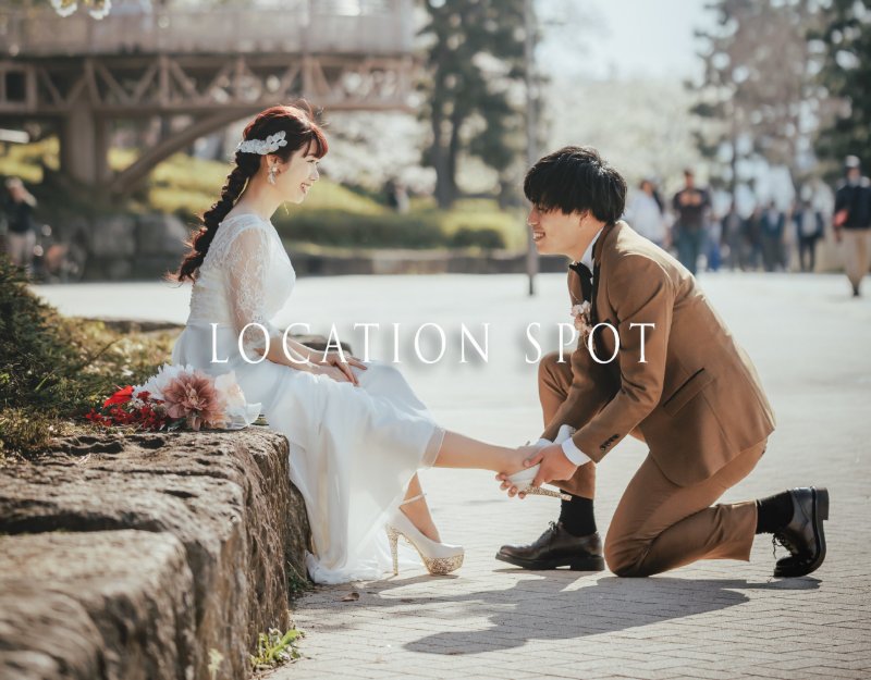 'Location Spot' banner: the groom kneels to adjust the bride's shoe beside cherry blossoms in a park.